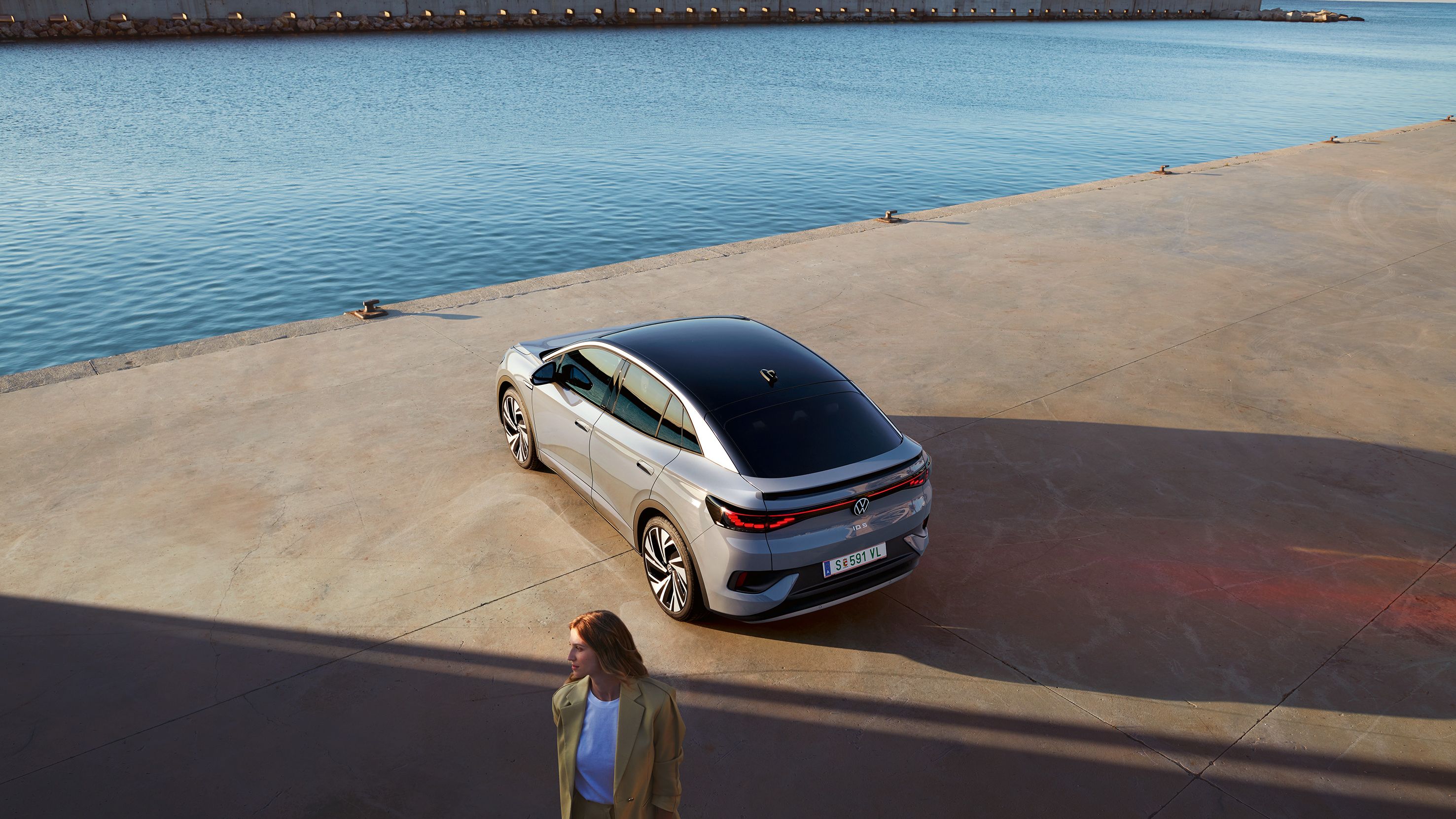 Vista superior oblíqua da traseira de um VW ID.5 no cais de um porto, em primeiro plano vê-se uma mulher