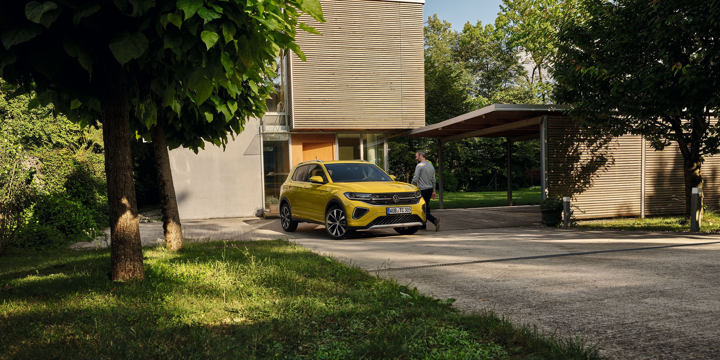 Um homem prepara-se para entrar num VW T-Cross amarelo estacionado na entrada do jardim de uma casa, vista frontal.