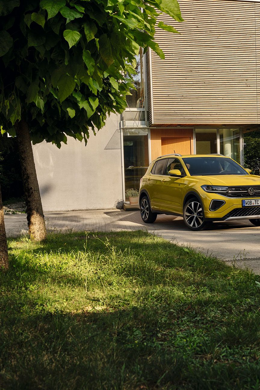 Um homem prepara-se para entrar num VW T-Cross amarelo estacionado na entrada do jardim de uma casa, vista frontal.