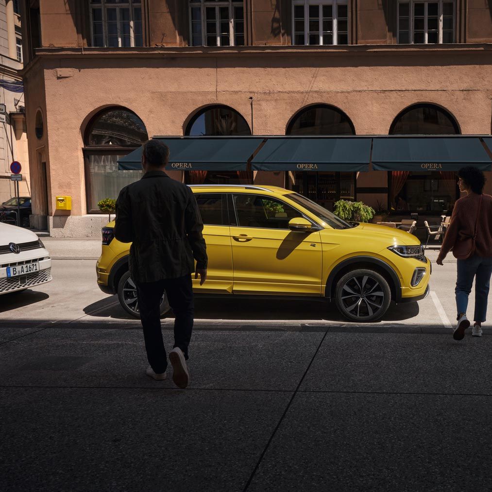 Um homem e uma mulher aproximam-se de um VW T-Cross amarelo na berma da estrada.