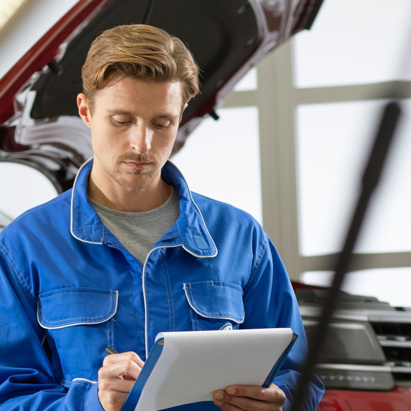 Empregado de serviços VW verifica uma lista de verificação de serviços em frente a um VW vermelho com capô aberto