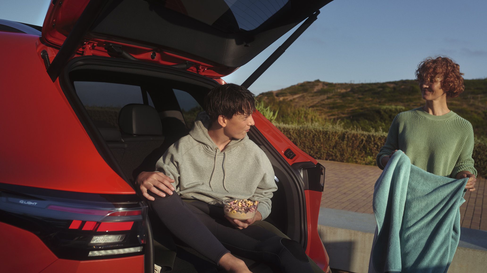 Fotografia da traseira de um VW Tiguan vermelho com a porta da bagageira aberta. Um jovem come descontraidamente os seus cereais na bagageira do veículo, uma jovem está à sua frente com uma toalha de praia.