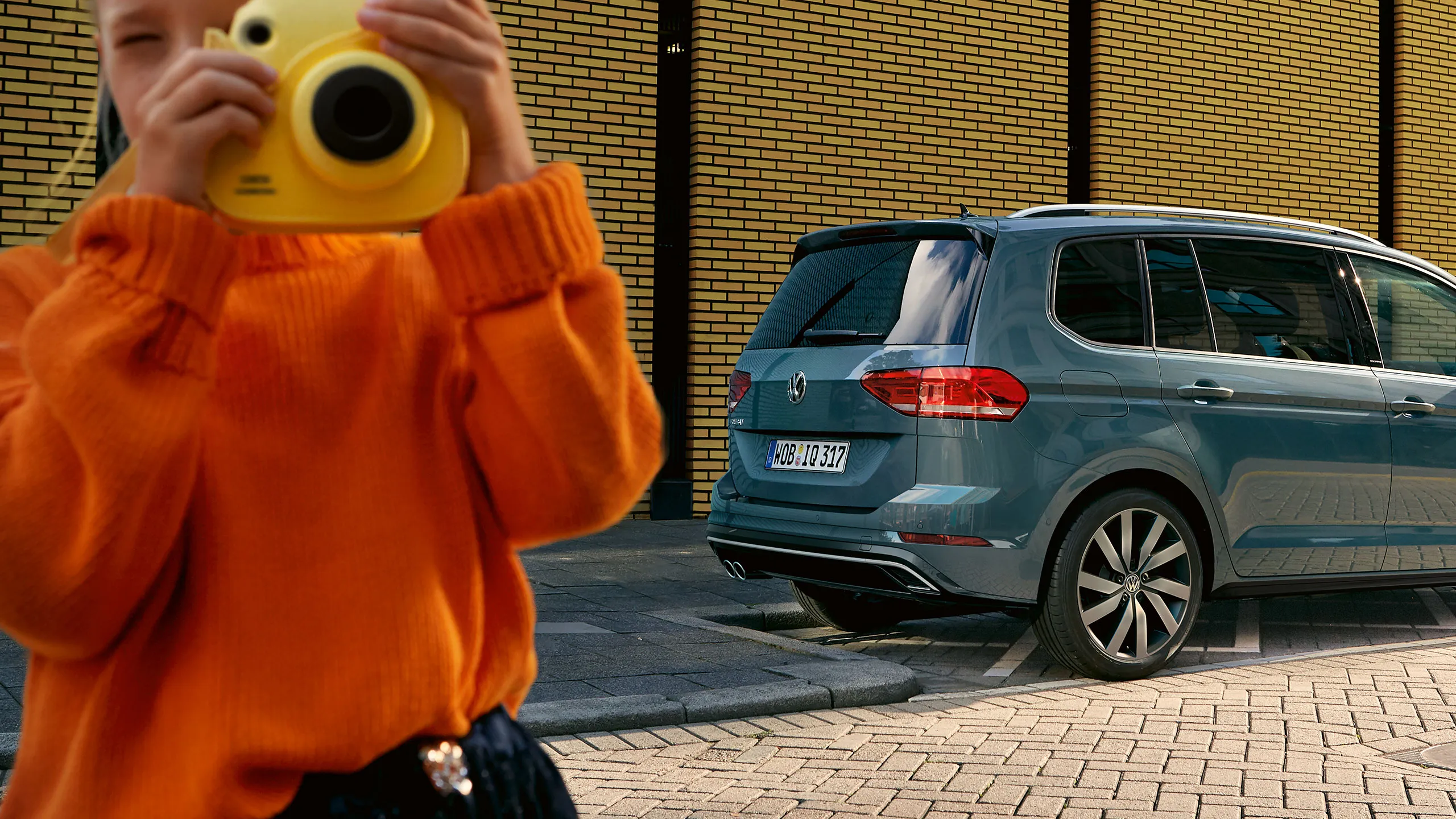 Uma criança com uma grande máquina fotográfica amarela fica em frente de um carro VW azul com pneus de Verão