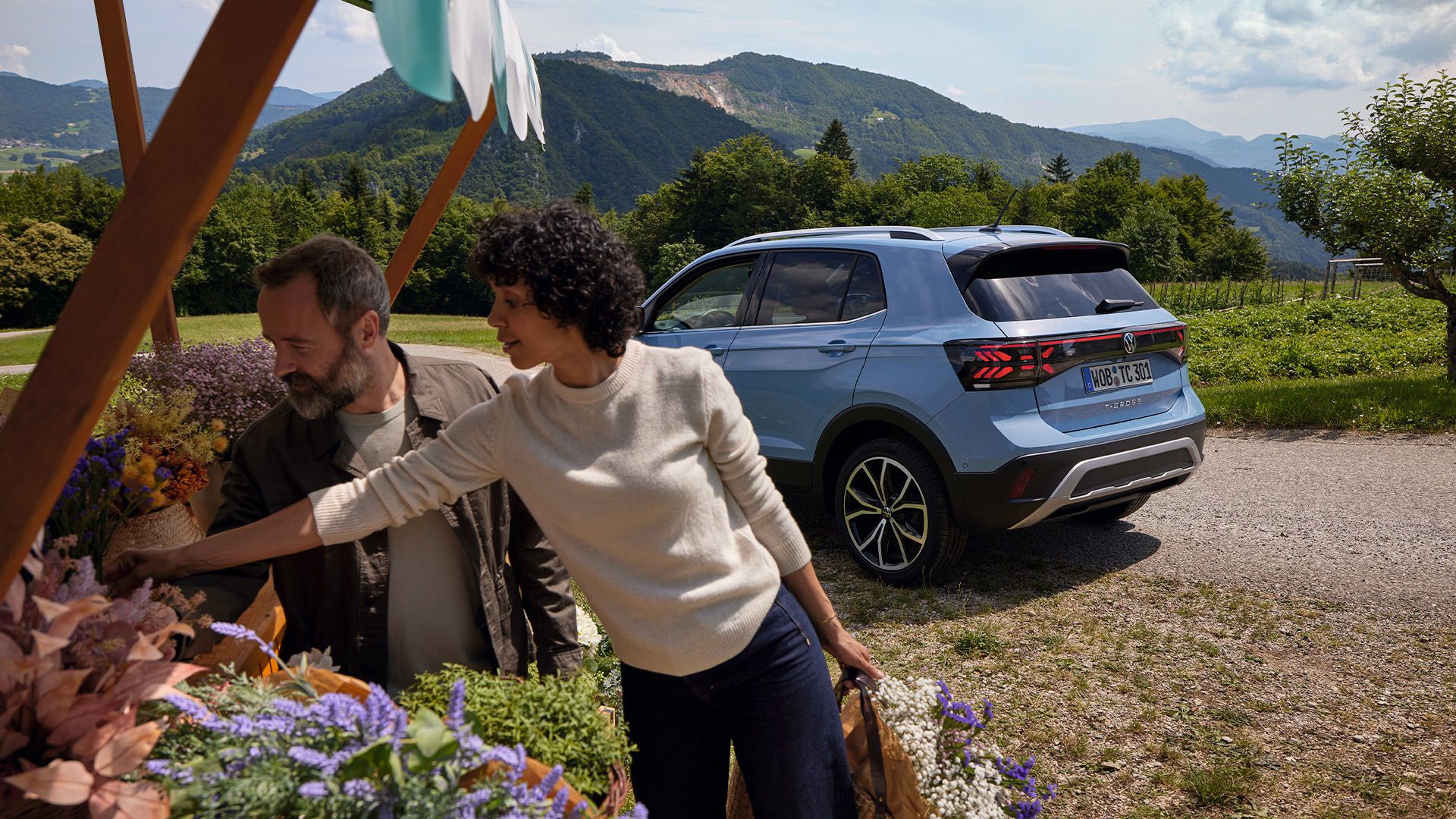 Um homem e uma mulher numa florista, atrás deles um VW T-Cross azul perante uma paisagem montanhosa.