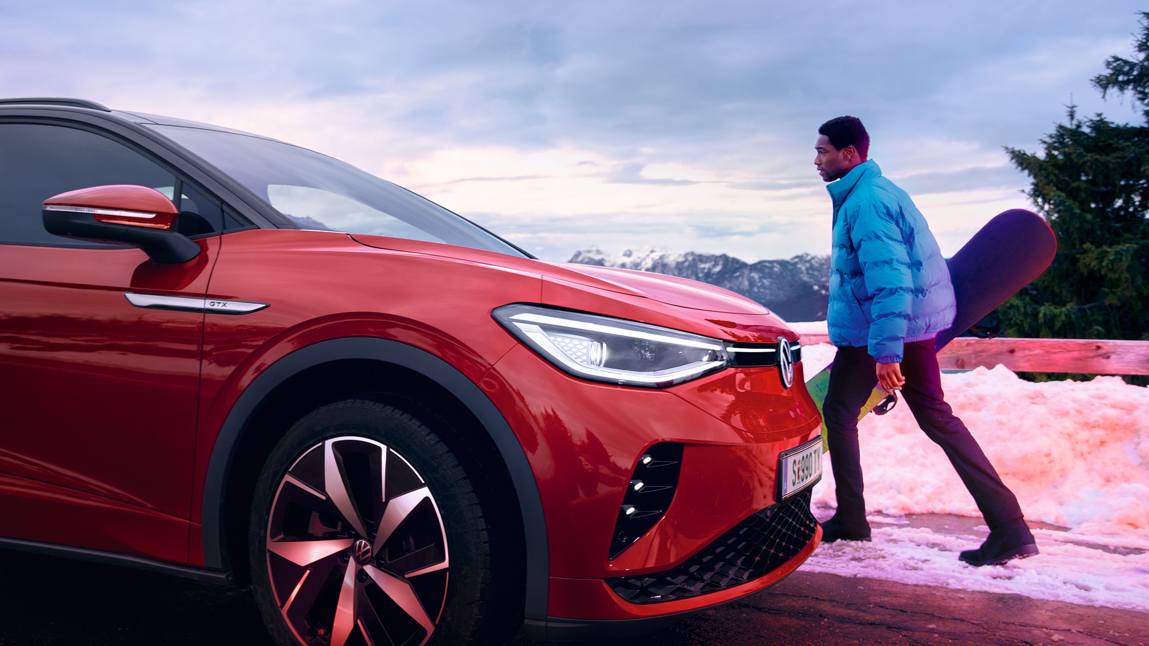 VW ID.4 GTX em vermelho de lado numa estrada na neve. Homem com casaco de Inverno e prancha de snowboard na mão caminha em direção ao SUV desportivo 100% elétrico. 