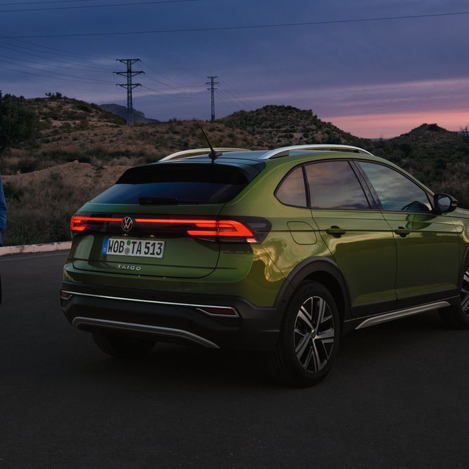 Vista lateral e traseira de VW Taigo verde estacionado, com as luzes acesas; homem em pé, atrás do automóvel