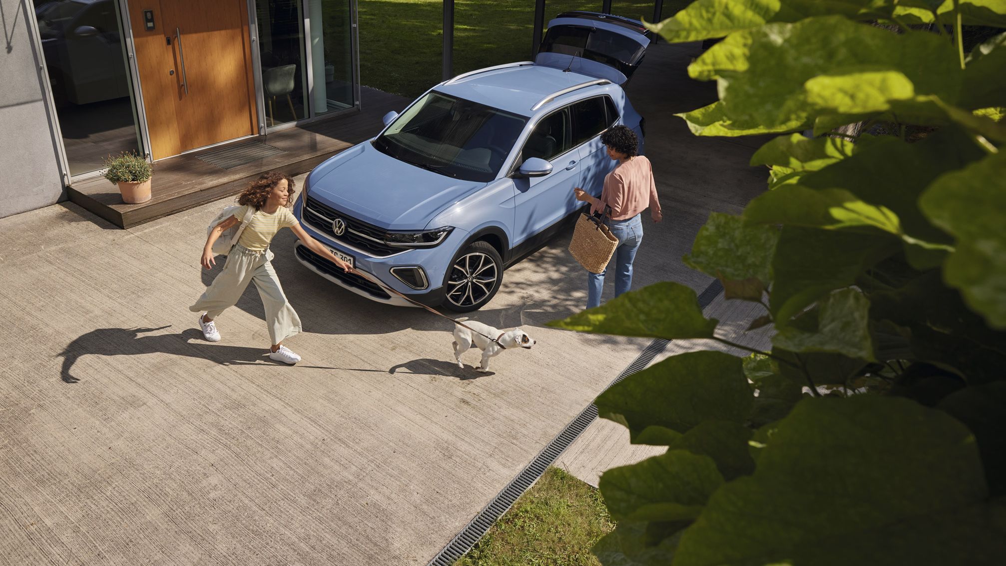 Uma mulher com uma cesta e uma menina com um cão perante um VW T-Cross azul com a bagageira aberta numa via de entrada.