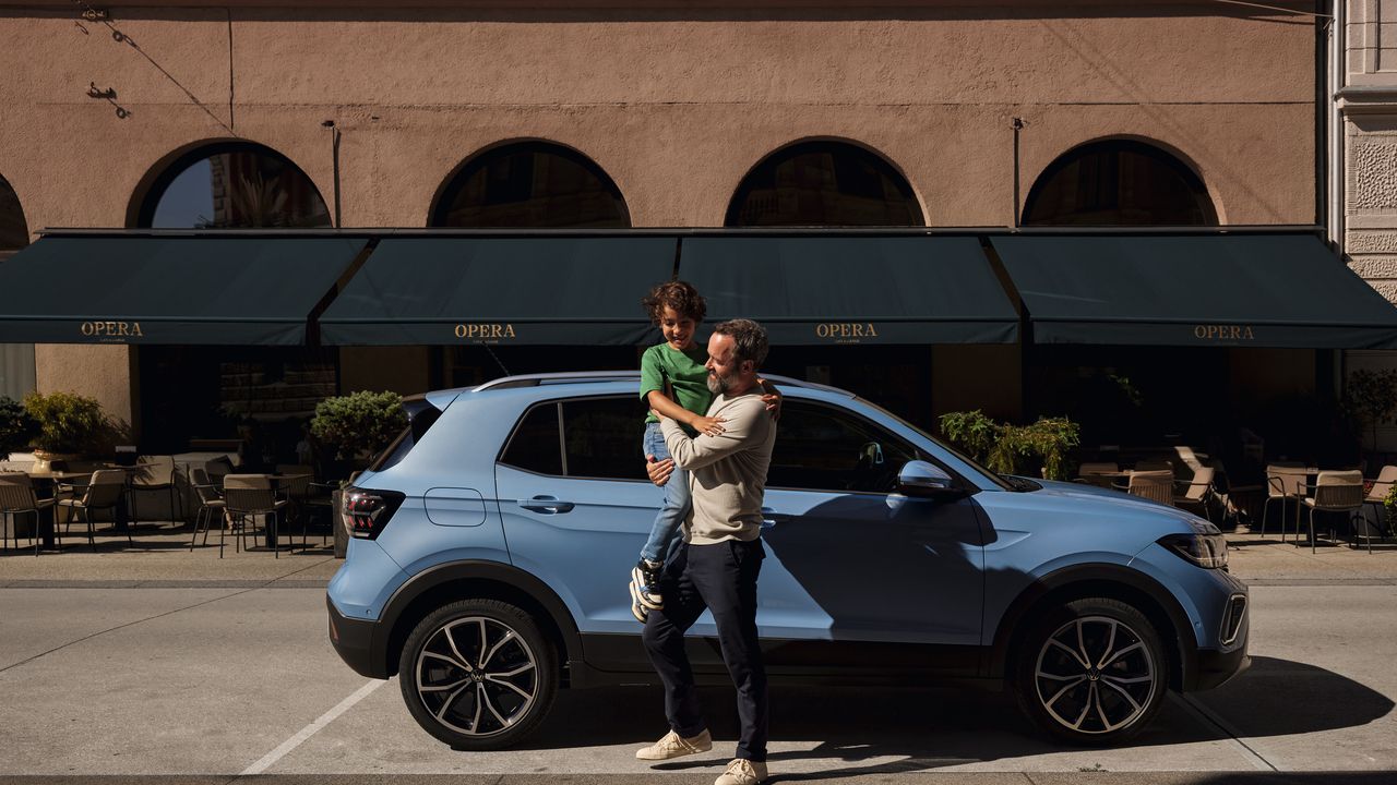 Um homem segura um menino alegre nos braços perante um VW T-Cross azul.