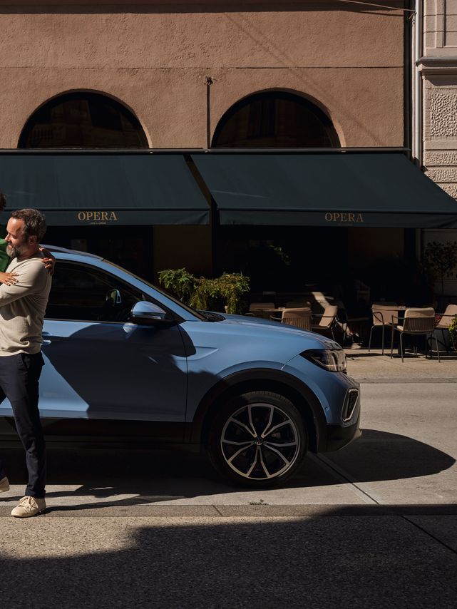 Um homem segura um menino alegre nos braços perante um VW T-Cross azul.