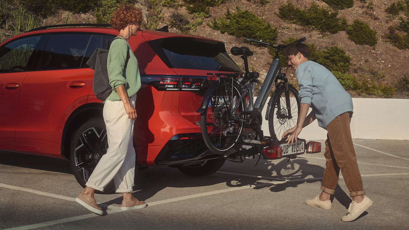 Fotografia da traseira de um VW Tiguan vermelho com dispositivo de engate de reboque opcional. Um casal prende uma bicicleta ao mesmo.