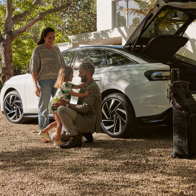 VW ID.7 estacionado à sombra, uma família carrega a bagageira. Em primeiro plano estão malas.