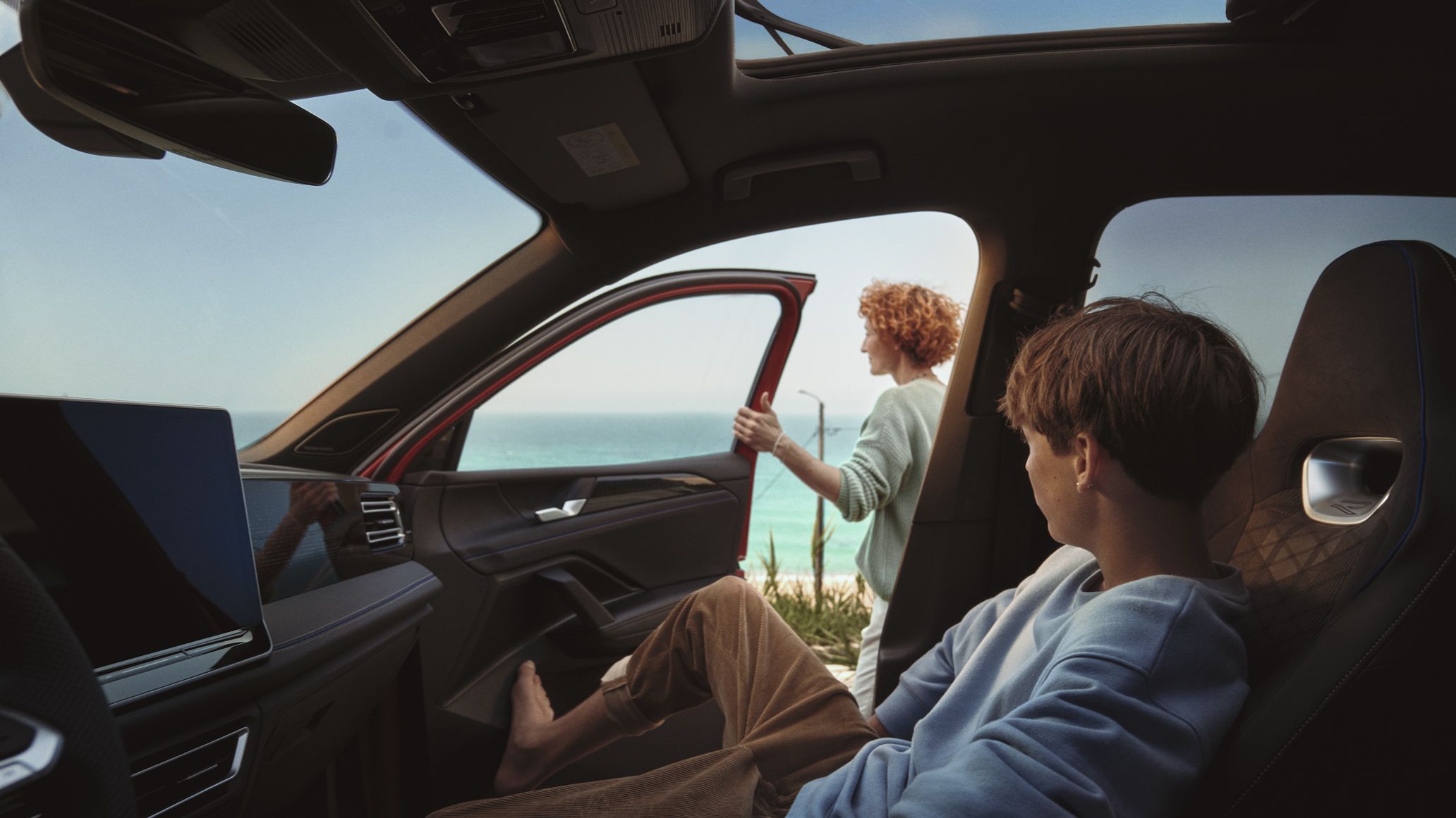 Vista do habitáculo do VW Tiguan. Um jovem está a descansar no banco do passageiro, uma mulher está de pé do lado de fora da porta aberta do passageiro.