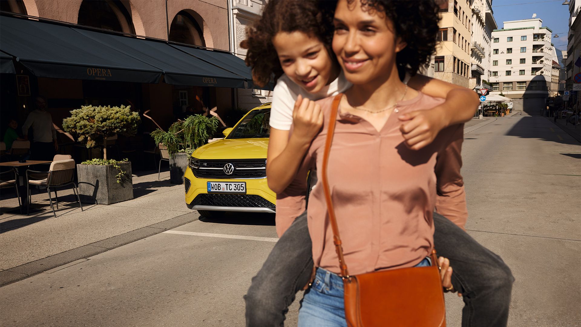 Numa rua no centro da cidade, uma mulher carrega uma menina às costas com um VW T-Cross amarelo em segundo plano.