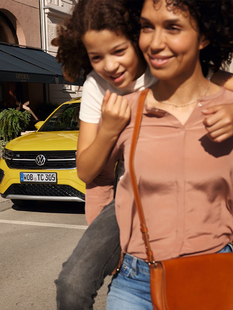 Numa rua no centro da cidade, uma mulher carrega uma menina às costas com um VW T-Cross amarelo em segundo plano.