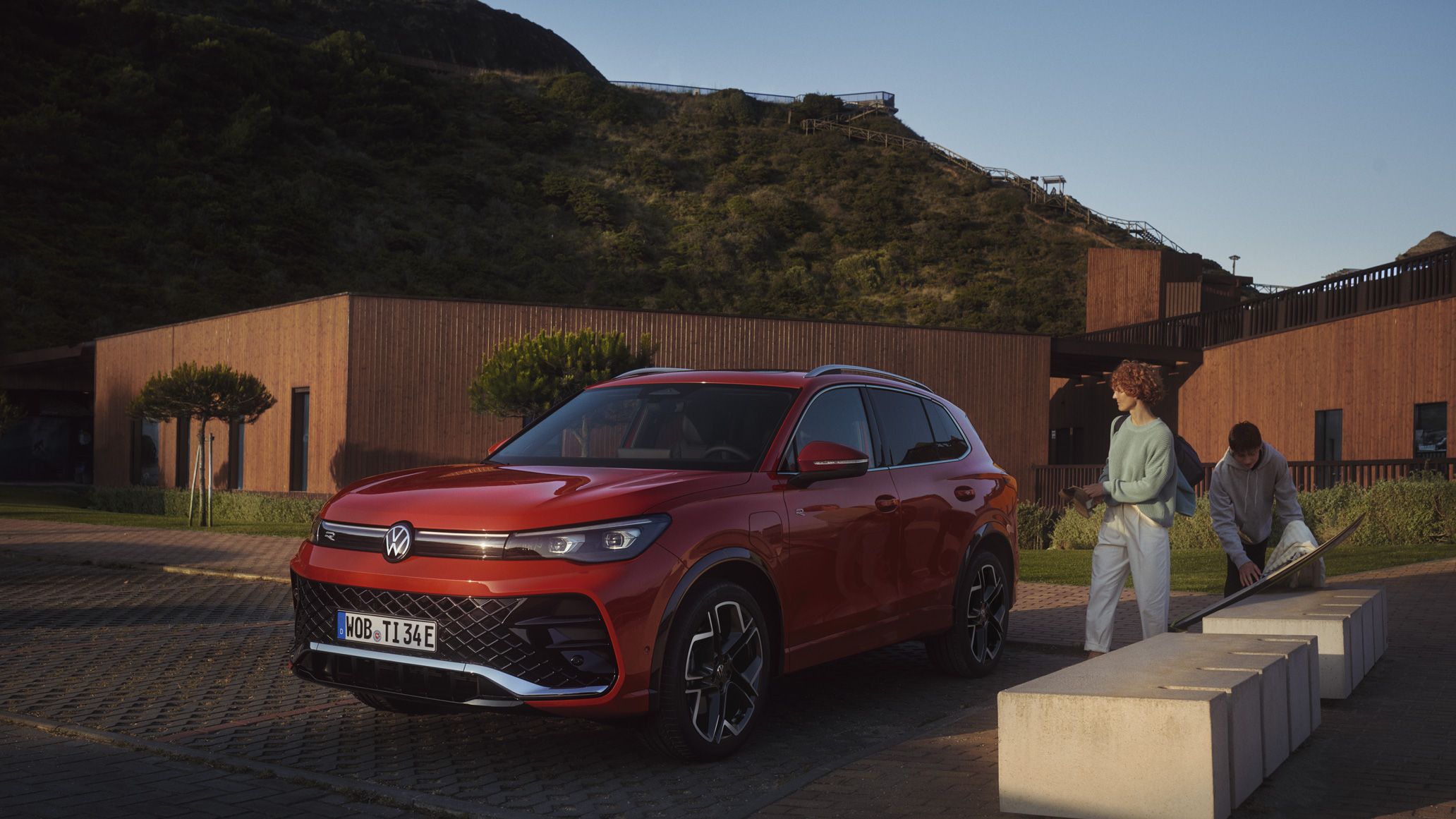 Vista frontal de um VW Tiguan vermelho num parque de estacionamento. Um casal com um skate caminha em direção ao veículo.