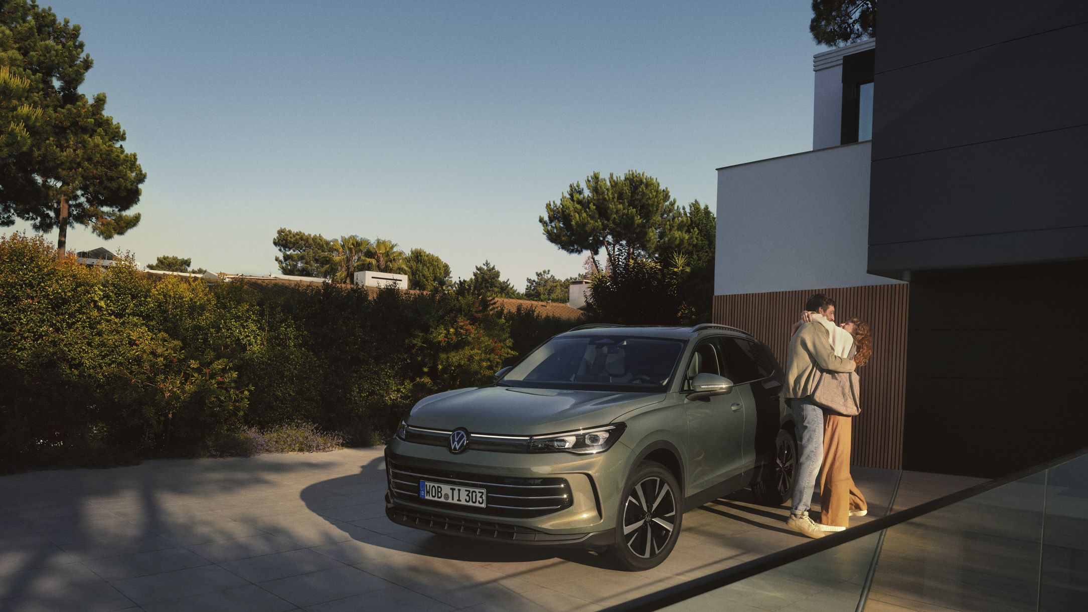 Vista frontal de um VW Tiguan verde estacionado na entrada de um edifício residencial moderno. Um jovem casal está ao lado dele e abraça-se.