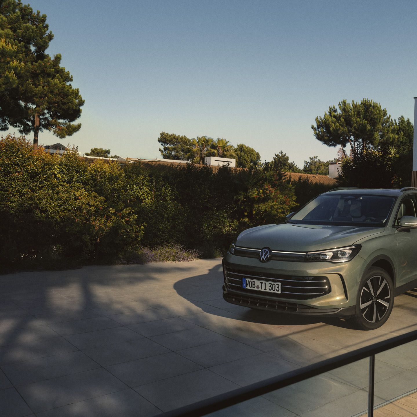 Vista frontal de um VW Tiguan verde estacionado na entrada de um edifício residencial moderno. Um jovem casal está ao lado dele e abraça-se.