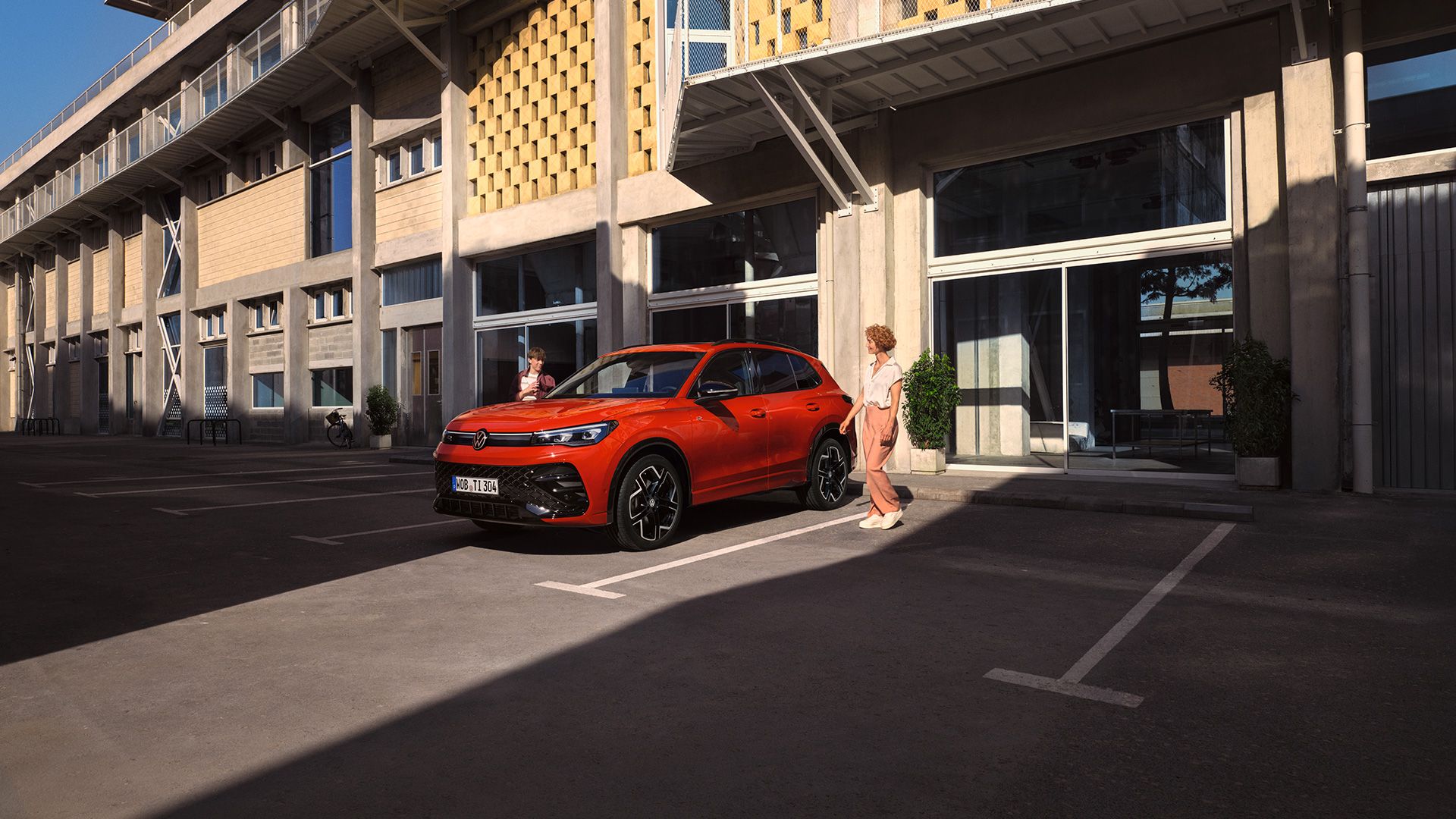 Vista lateral de um VW Tiguan vermelho, parado em frente a um edifício, uma jovem passa do lado do condutor.