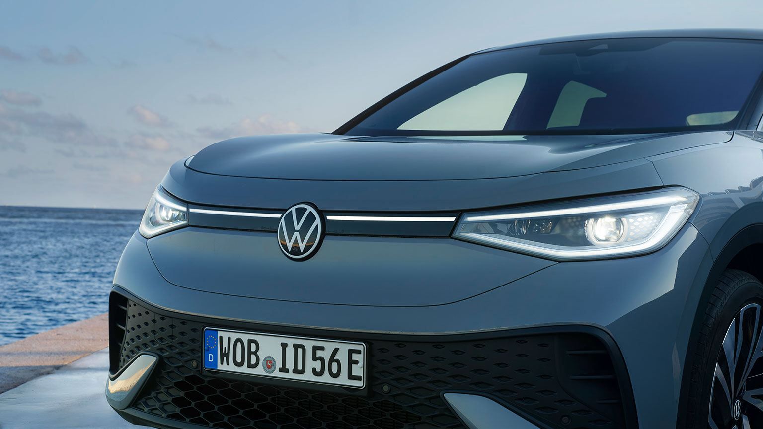 Dianteira de um VW ID.5 parado no cais de um porto ao entardecer, faróis LED acesos