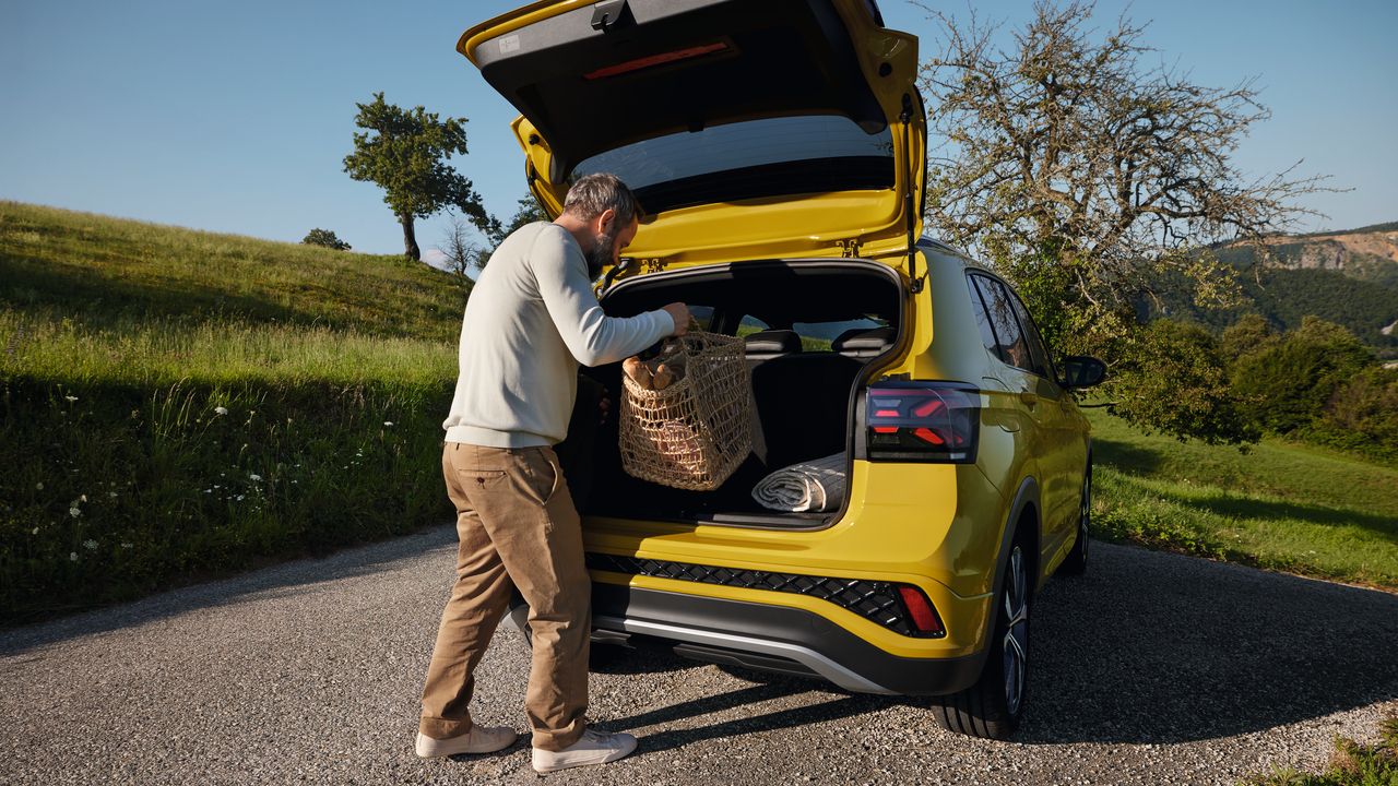 Um homem coloca uma grande cesta de piquenique na bagageira de um VW T-Cross amarelo que se encontra numa estrada com paisagem montanhosa.