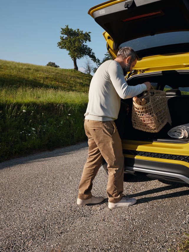 Um homem coloca uma grande cesta de piquenique na bagageira de um VW T-Cross amarelo que se encontra numa estrada com paisagem montanhosa.