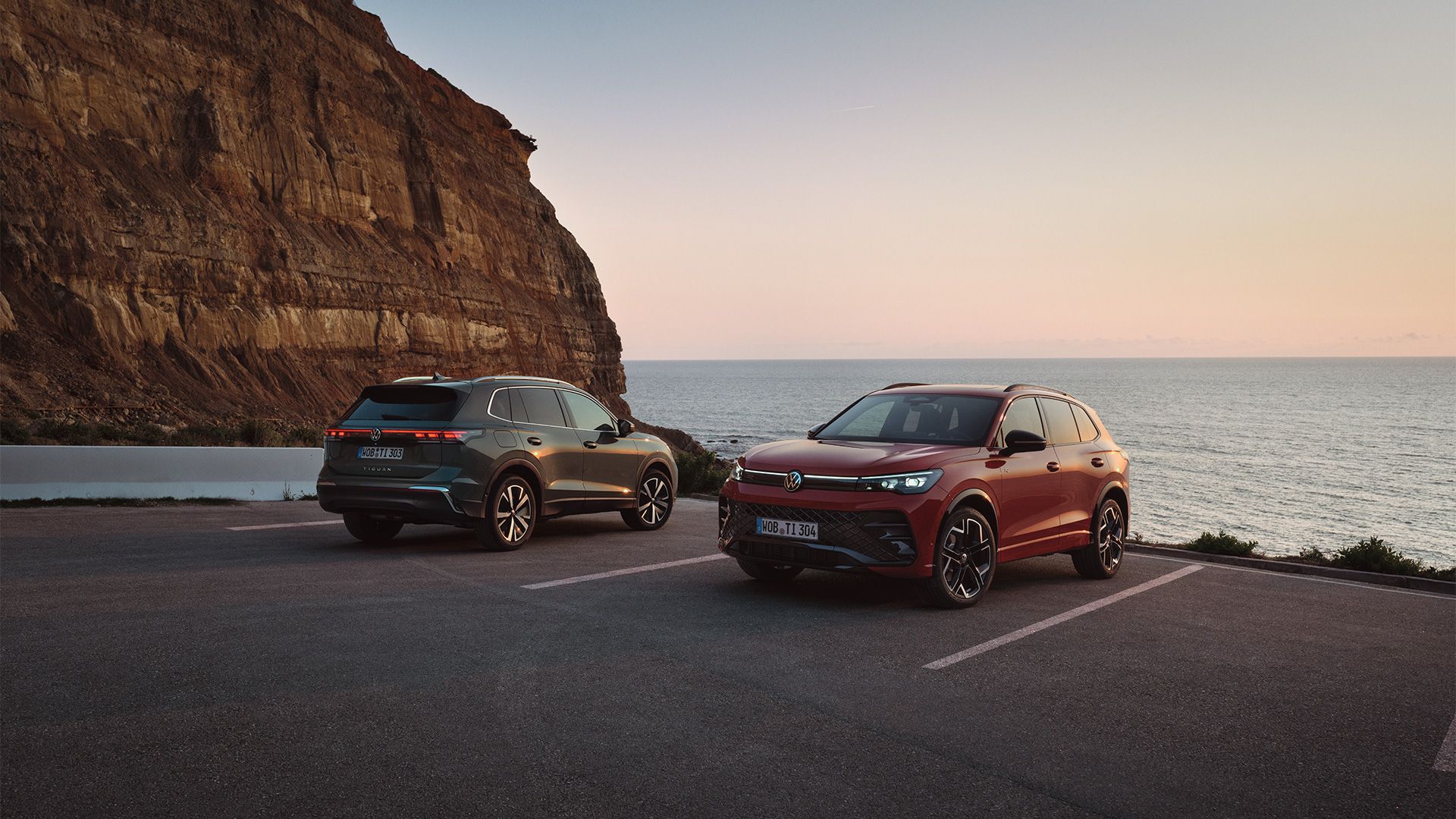 Dois VW Tiguan verde escuro e vermelho estacionados em frente a uma paisagem costeira rochosa.