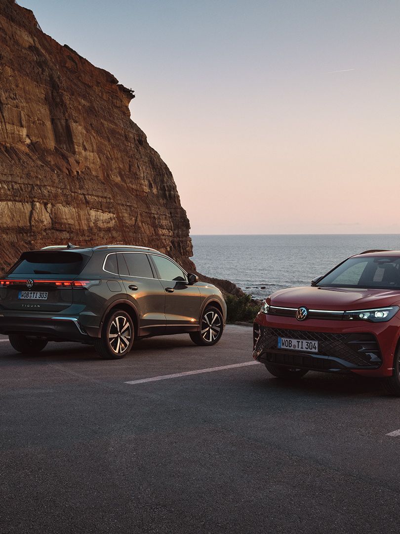 Dois VW Tiguan verde escuro e vermelho estacionados em frente a uma paisagem costeira rochosa.