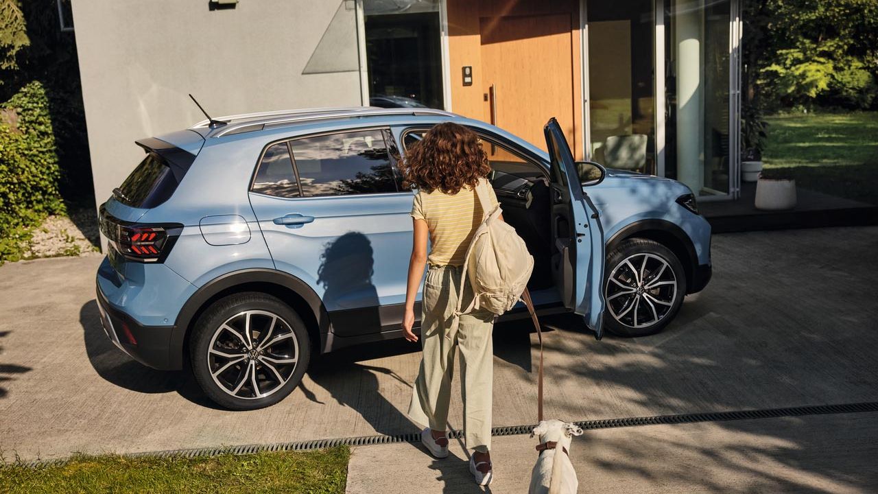 Uma menina com um cão aproxima-se de um VW T-Cross azul numa entrada. A porta do passageiro está aberta.