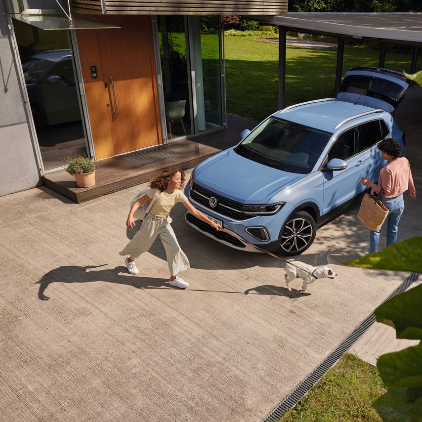 Uma mulher com uma cesta e uma menina com um cão perante um VW T-Cross azul com a bagageira aberta numa via de entrada.