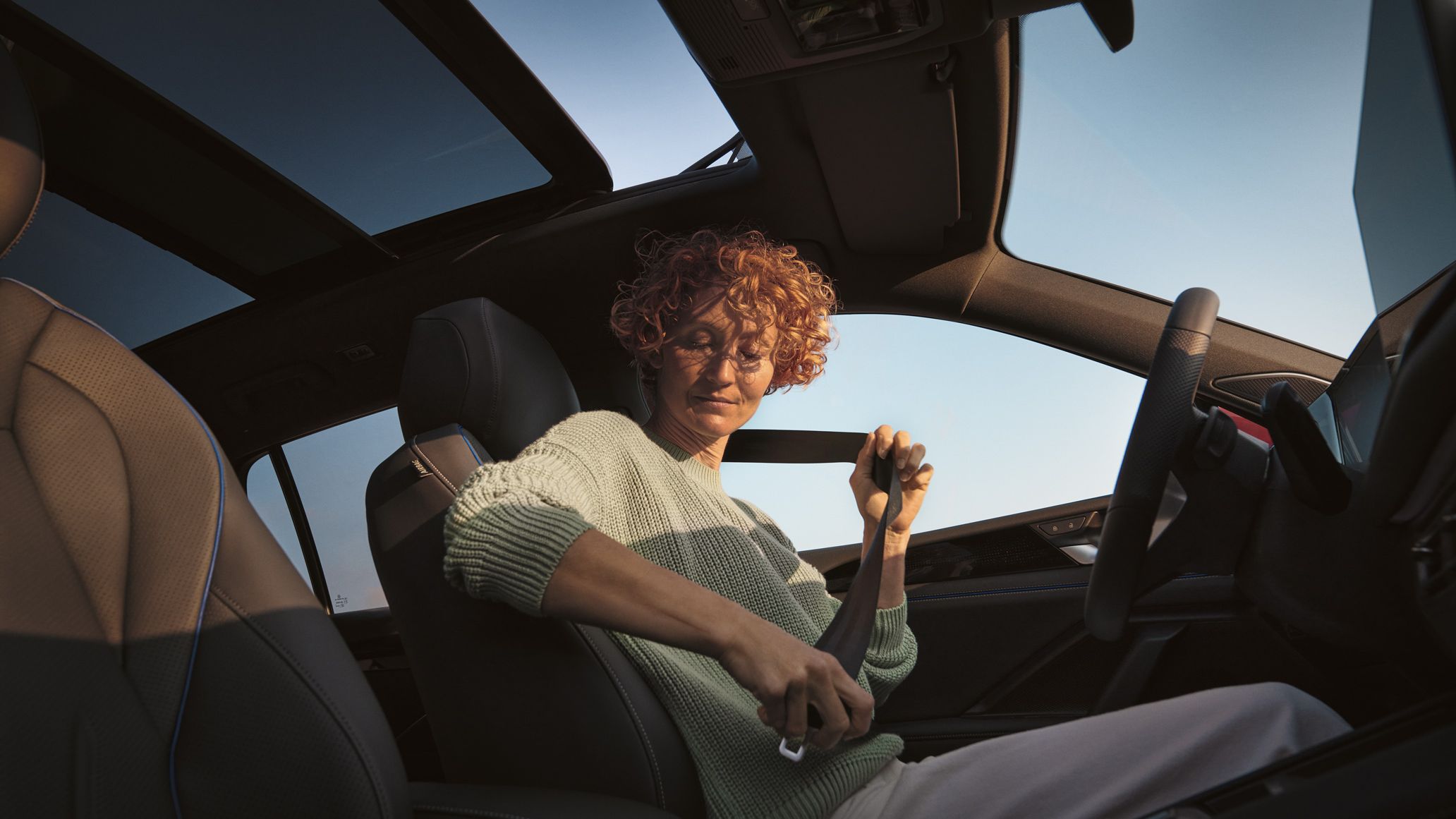 Fotografia do interior de um VW Tiguan com vista para o céu através do teto de abrir/correr panorâmico opcional semiaberto. Uma jovem tem o cinto de segurança colocado no banco do condutor.