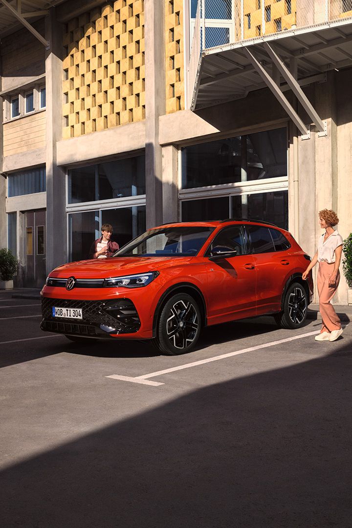 Vista lateral de um VW Tiguan vermelho, parado em frente a um edifício, uma jovem passa do lado do condutor.