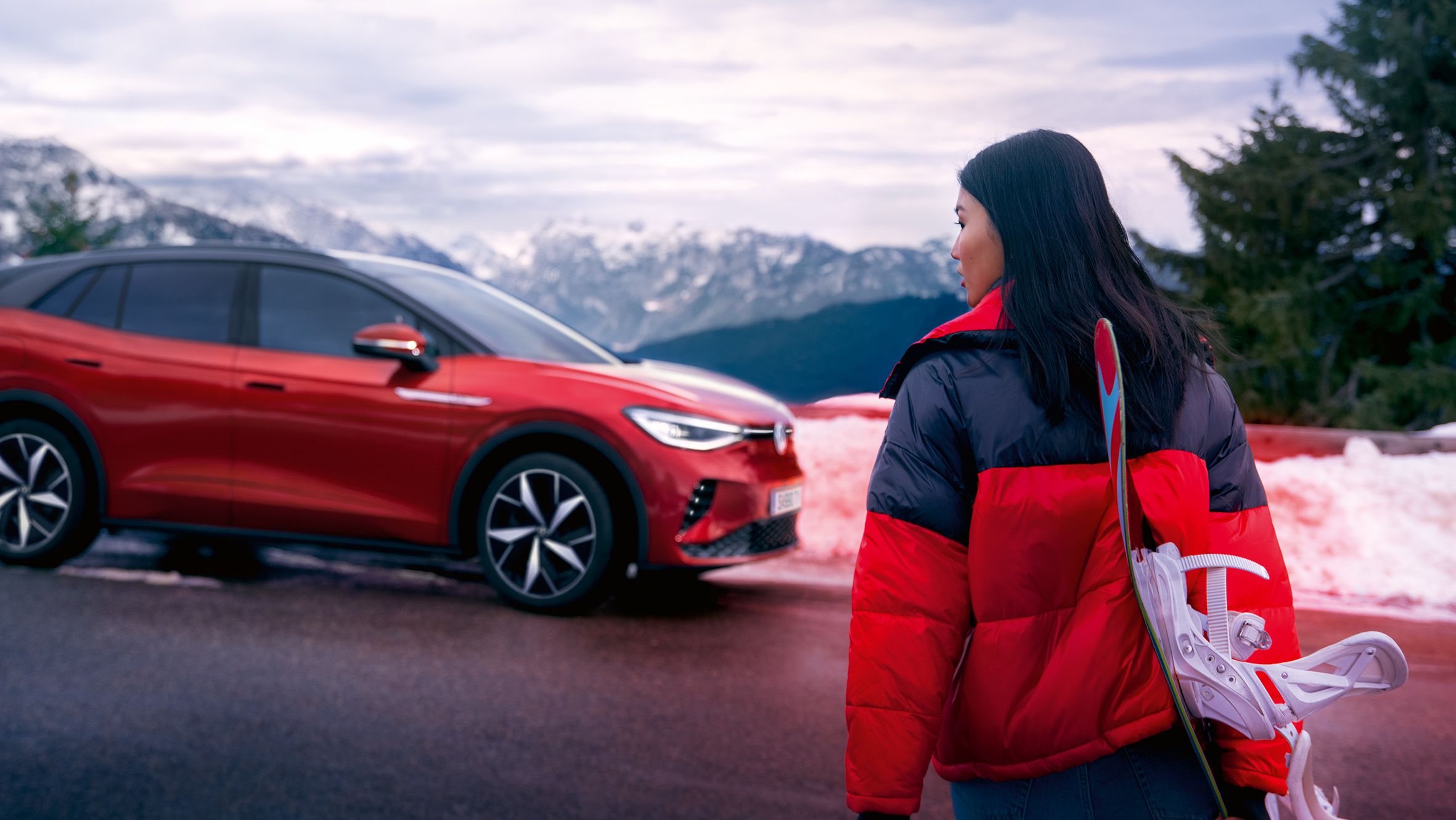 VW ID.4 GTX em vermelho na estrada numa paisagem de montanhas com neve. Mulher de casaco vermelho e preto atravessa a estrada com esquis e olha para a esquerda.