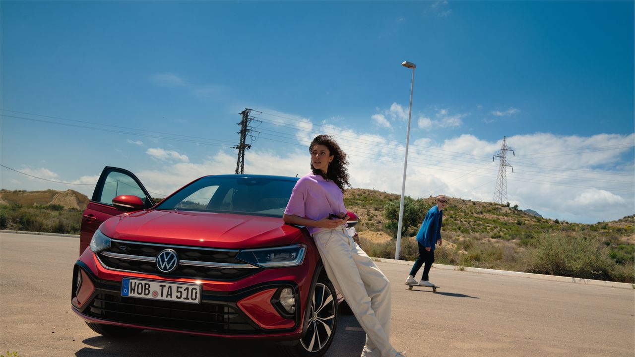 VW Taigo R-Line vermelho; vista de frente num parque de estacionamento. Mulher inclinada sobre o capot; homem passa de Skateboard