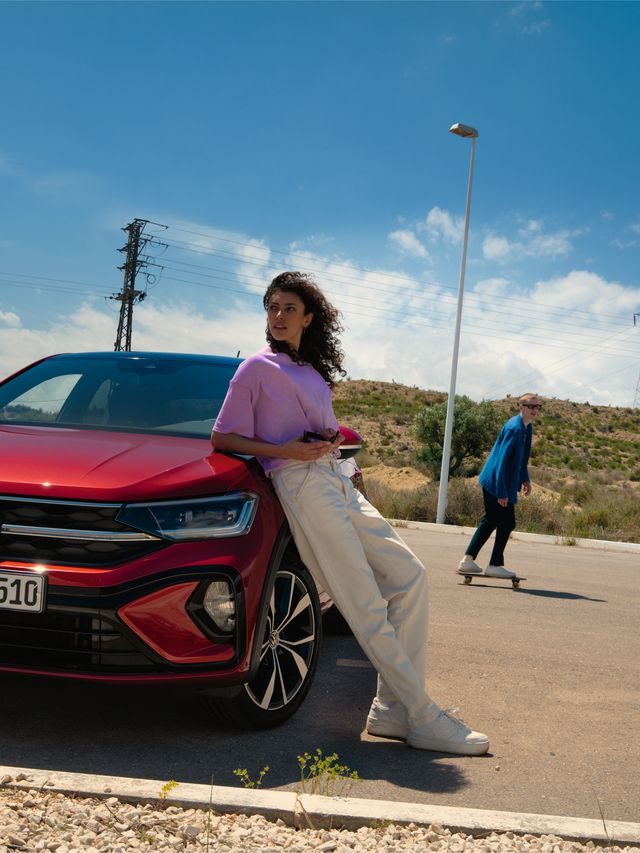 VW Taigo R-Line vermelho; vista de frente num parque de estacionamento. Mulher inclinada sobre o capot; homem passa de Skateboard